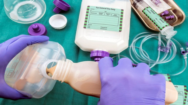 Person in purple gloves pouring beige liquid into a labeled medical container surrounded by syringes, tubing, and supplies on a green cloth.